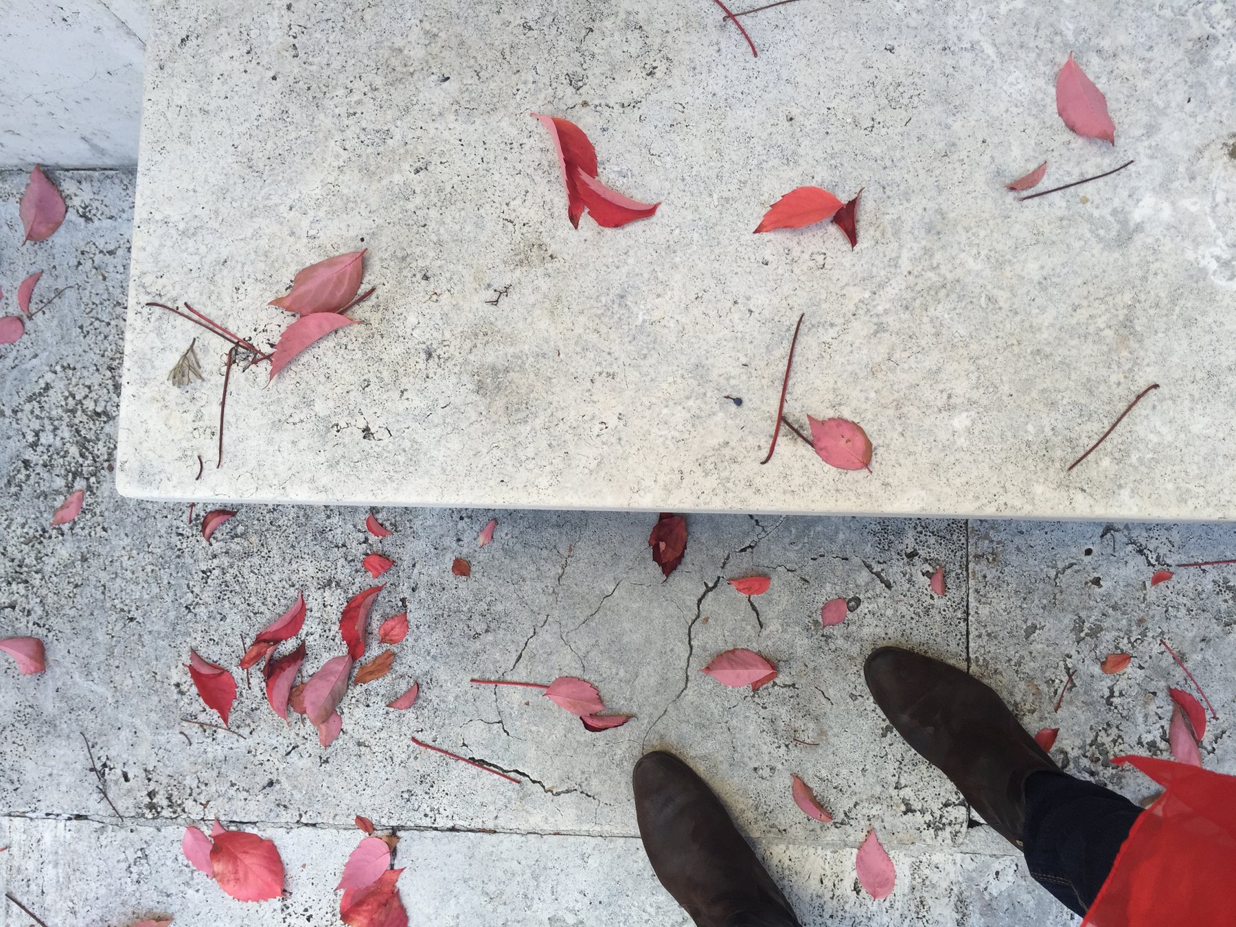 Looking straight down at the marble bench and surrounding stone paving, red leaves scattered across the surface, a pair of brown boots visible at the lower edge of the frame.