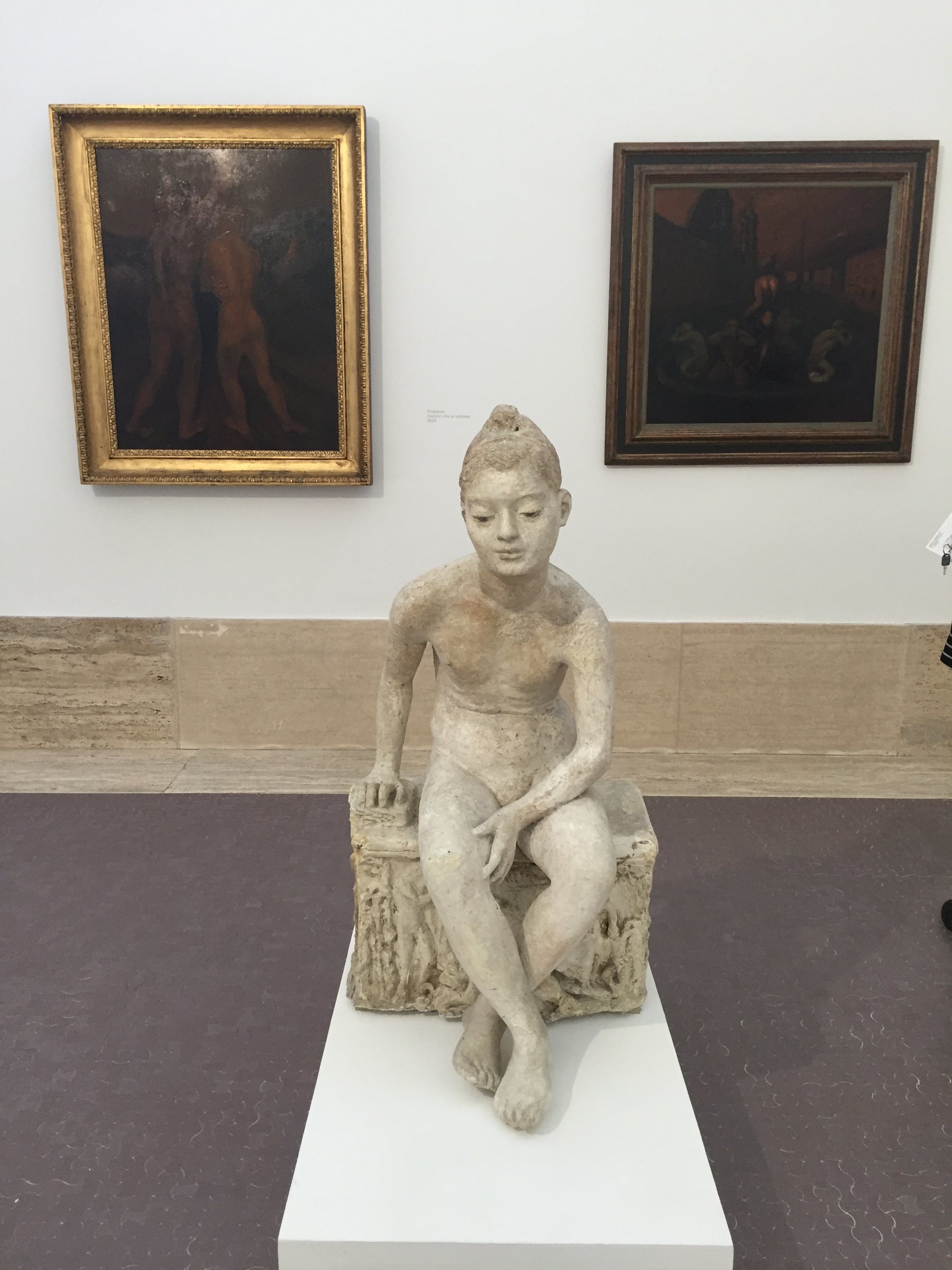 Interior of a gallery room, a pale plaster sculpture of a seated figure on a low plinth in the foreground, two dark oil paintings in ornate frames on the white wall behind, the figure gazing calmly downward.