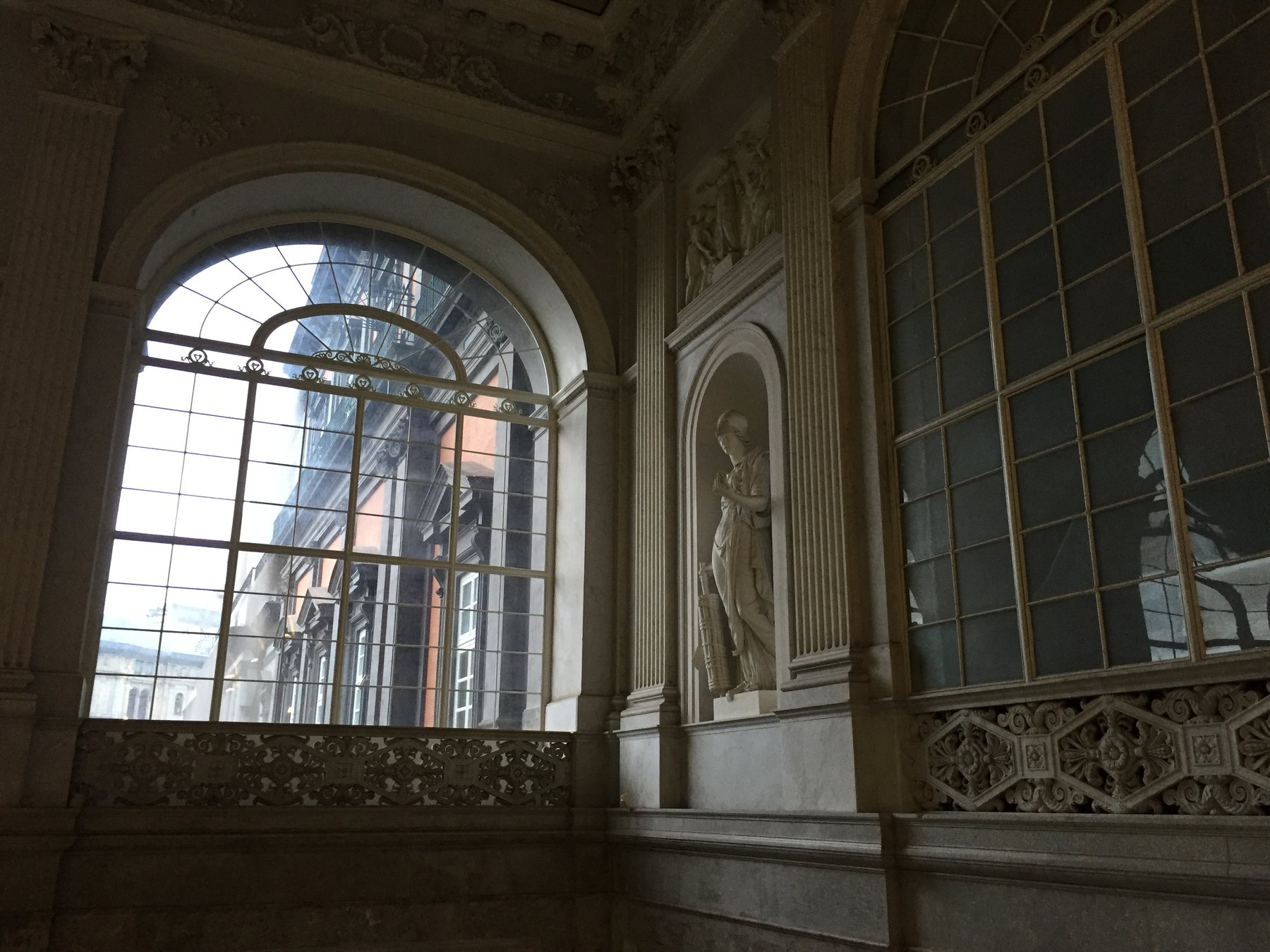 Interior view of the museum stairwell landing, a large arched window with iron grille letting in bright morning light, a classical marble statue set into a niche in the wall, ornate carved detailing throughout.