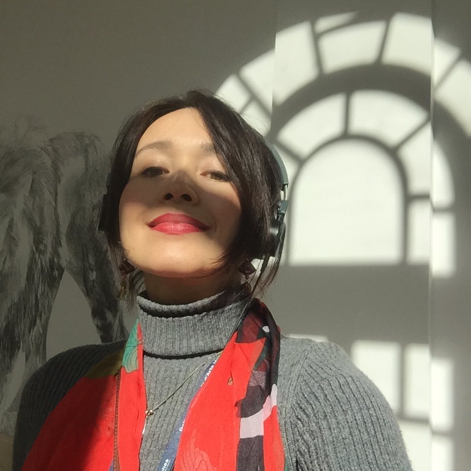 A woman with headphones inside the museum, face turned upward toward warm sunlight, the shadow of a large arched window cast on the wall behind her.