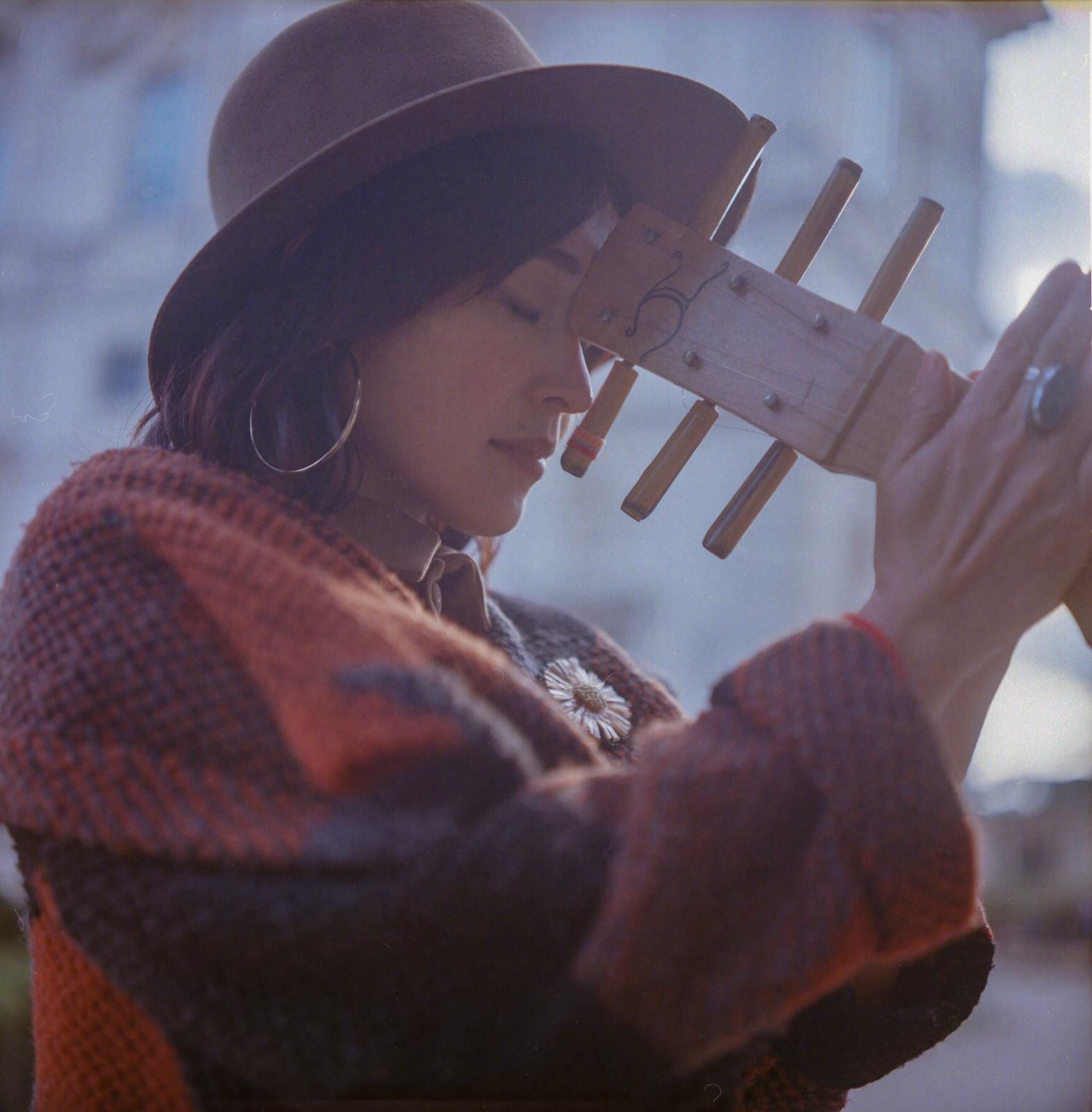 Colour film photograph of Jacelyn holding the sape neck upward to her forehead, eyes closed, hat, warm winter tones, analogue grain.