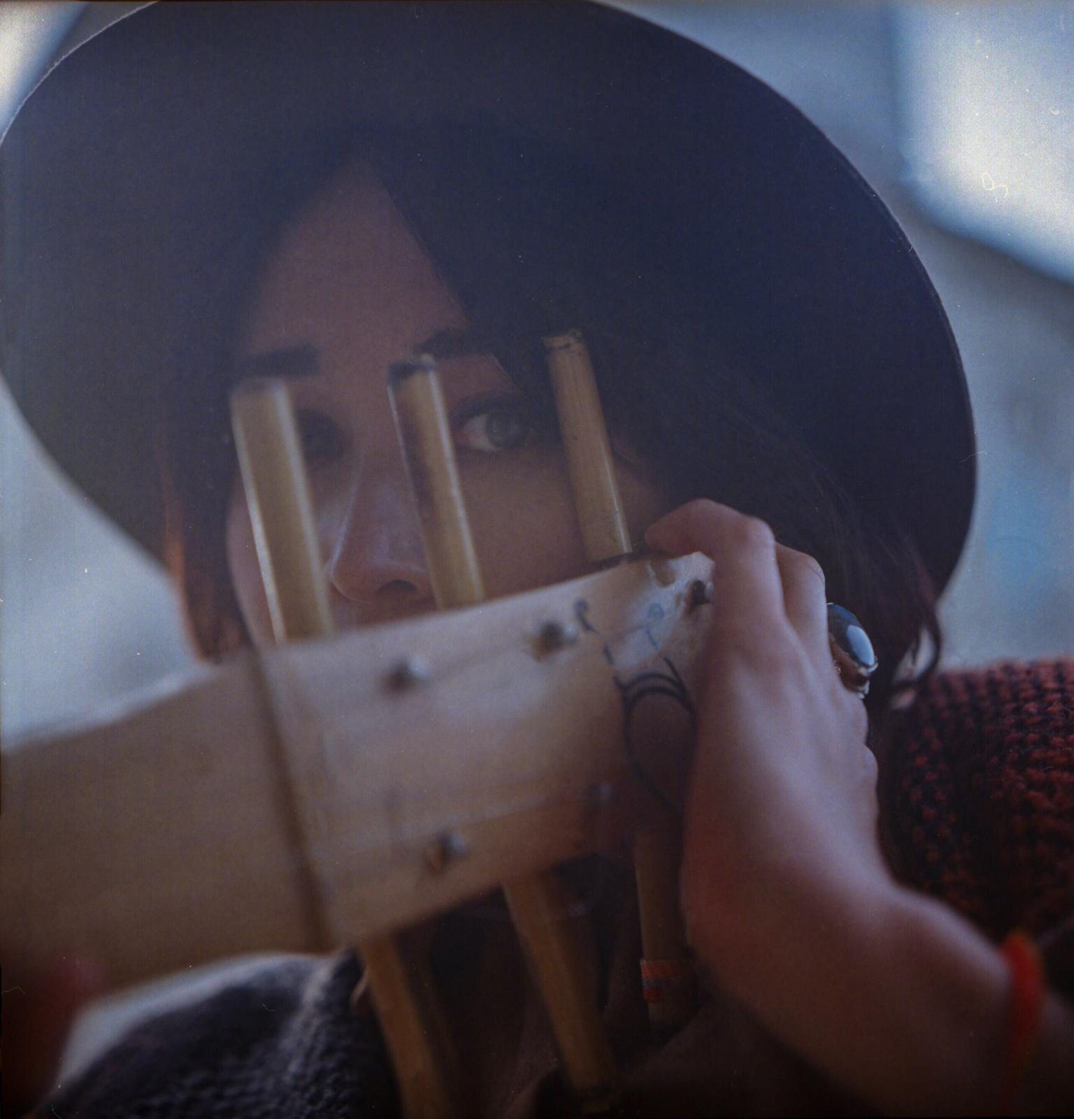 Colour film photograph of Jacelyn looking through the tuning pegs of the sape headstock held in front of her face, eyes visible between the wooden pegs, concentrated expression.