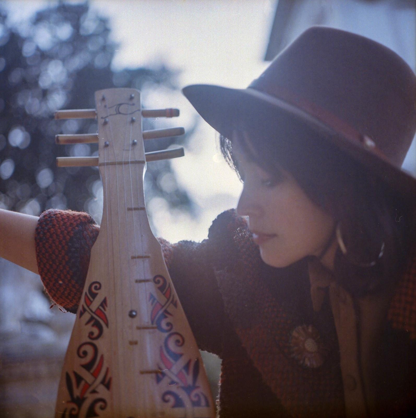 Colour film photograph of Jacelyn leaning toward the sape held upright, face close to the decorated body, knitted gloves, hat, winter garden out of focus behind.