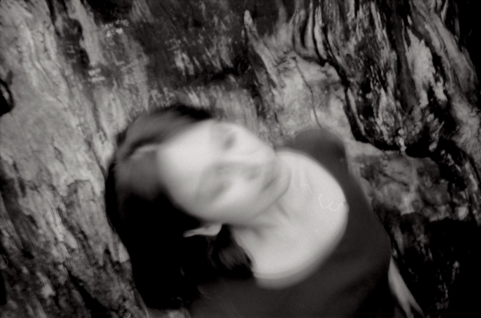Black and white photograph, blurred and dreamlike, of Jacelyn's face against the textured bark inside a tree hollow, film grain, abstract quality.