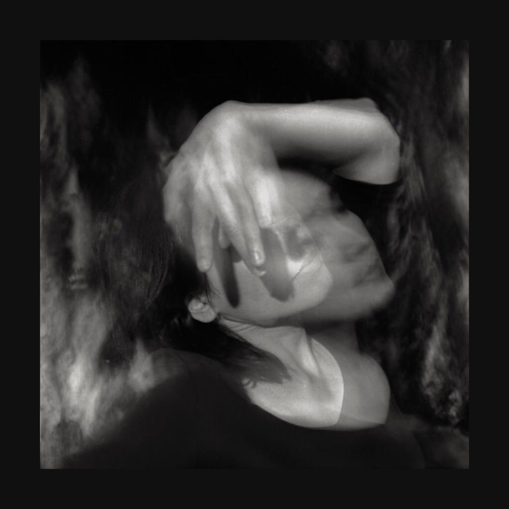 Black and white abstract photograph, hands covering a face, dreamlike and blurred, dark background with light catching the fingers and cheekbone, taken inside a tree hollow.