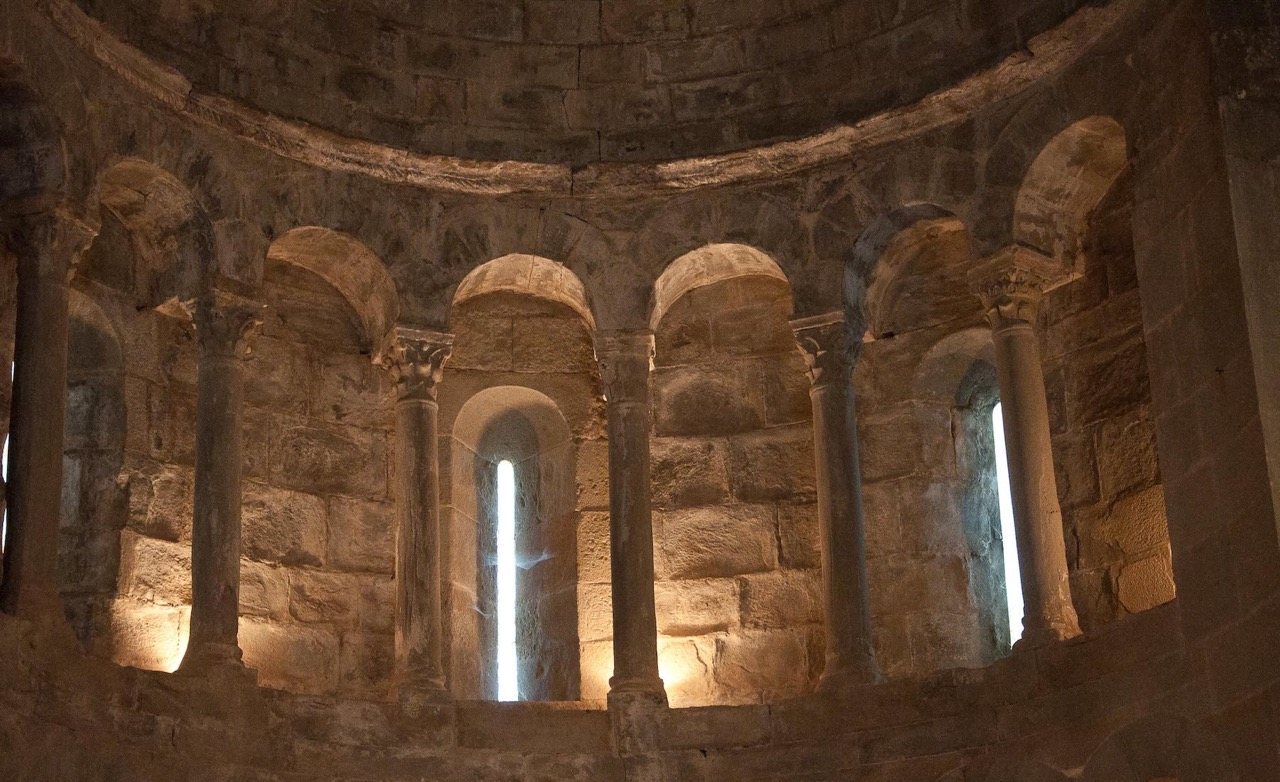 The illuminated stone apse of the church, narrow slit windows admitting light between columns.